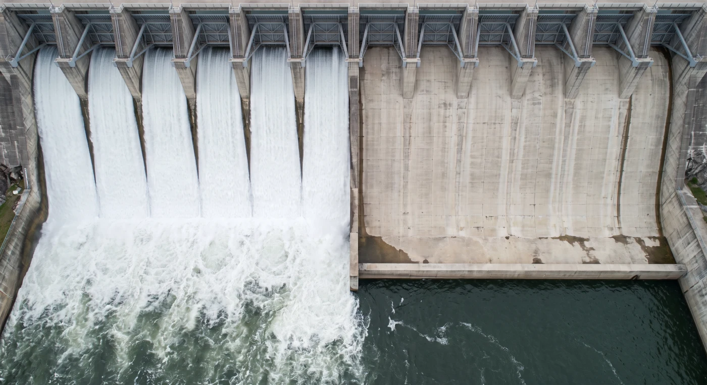A perpendicular aerial view of a dam spillway with twelve identical gates in a row: the six on the left open and discharging sheets of water, the six on the right closed with the concrete dry